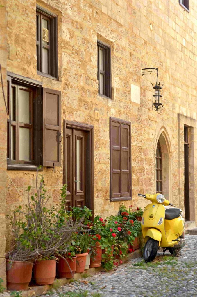 A charming street scene in Rhodes, Greece, featuring a quaint building, a yellow Vespa, and blooming flowers.