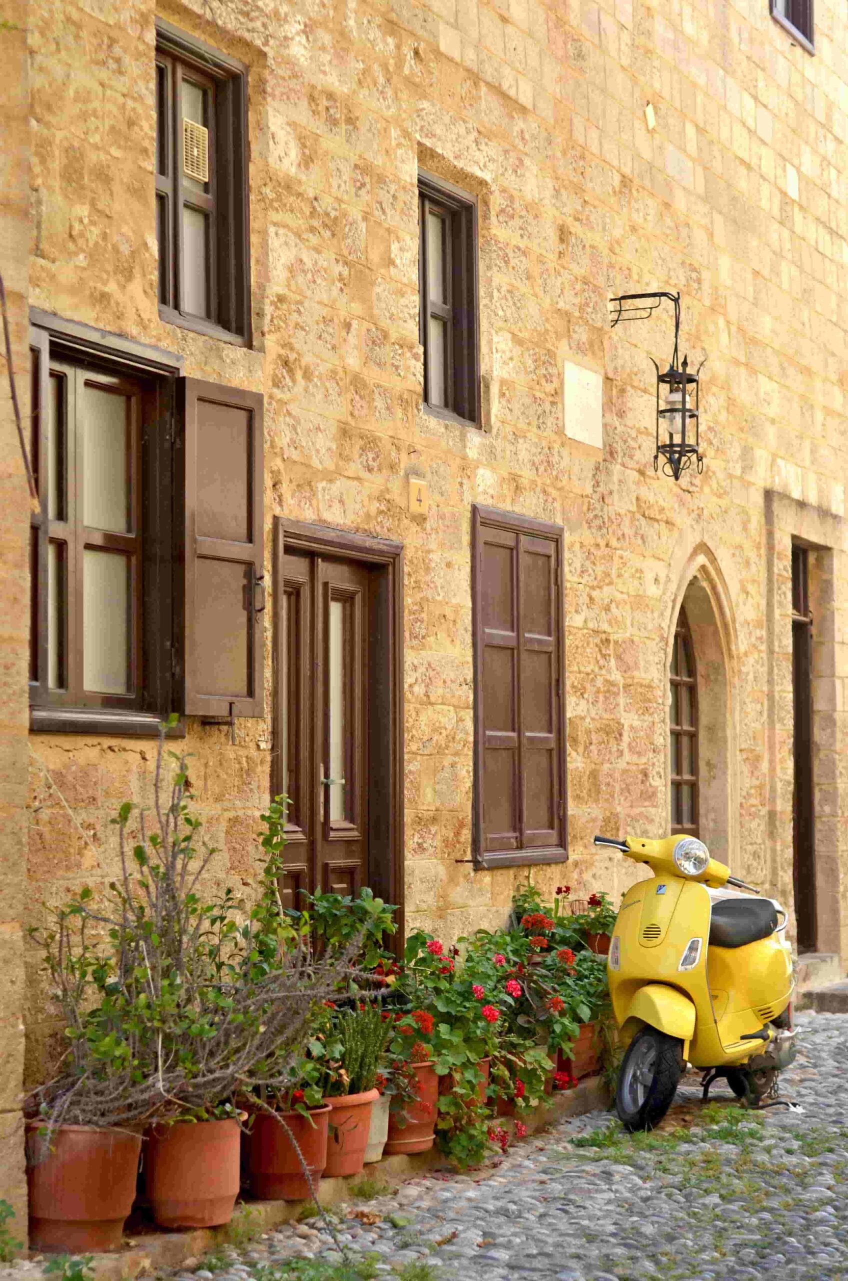 A charming street scene in Rhodes, Greece, featuring a quaint building, a yellow Vespa, and blooming flowers.